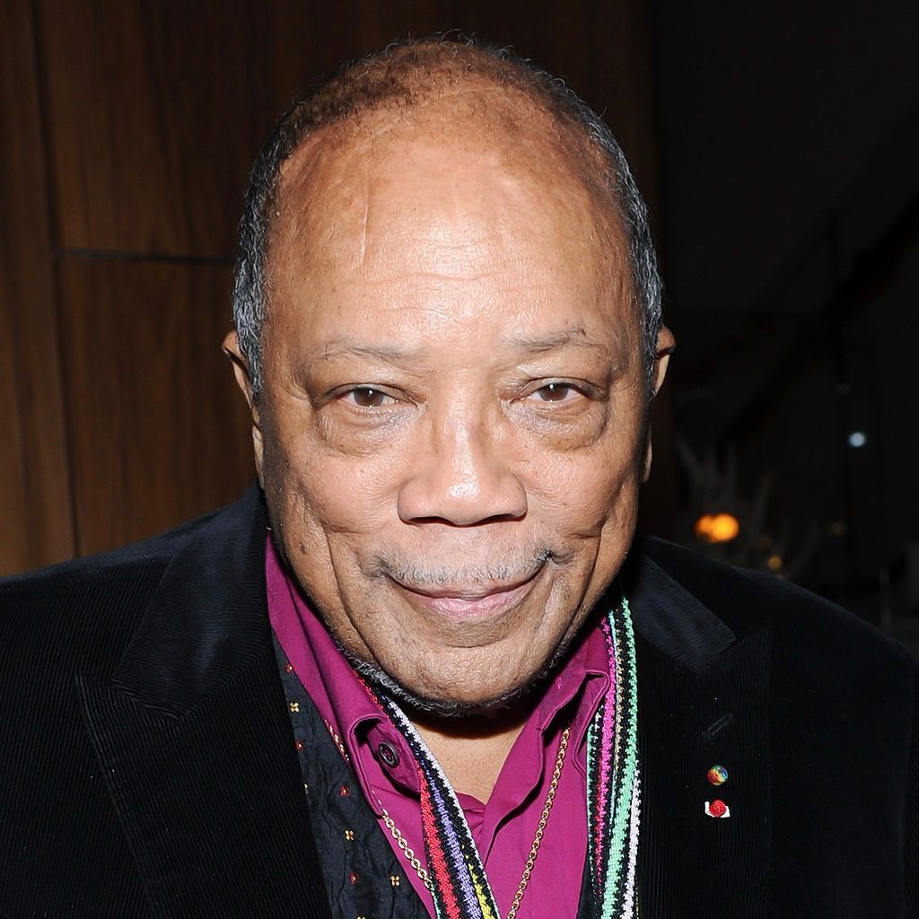 Quincy Jones wearing a dark blazer over a pink shirt, a patterned scarf, and colorful beaded necklaces.