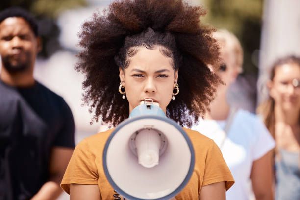 A person with curly hair holds a megaphone in front of their mouth, surrounded by others, conveying a message or protest.