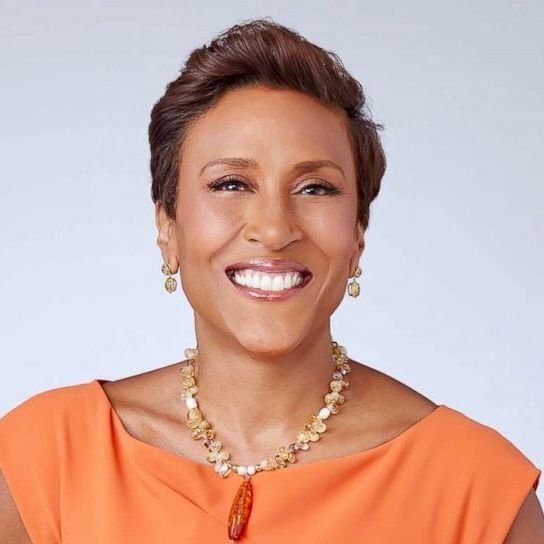 Robin Roberts smiling at the camera, wearing an orange top and a beaded necklace with a large, amber-colored pendant.