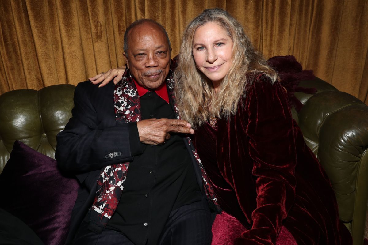 Quincy Jones and Barbra Streisand sit side-by-side on a green leather couch in front of gold curtains.