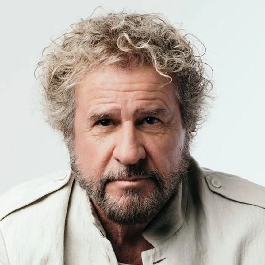 A person with curly hair and a beard, wearing a light-colored jacket, looking directly at the camera against a grey background.
