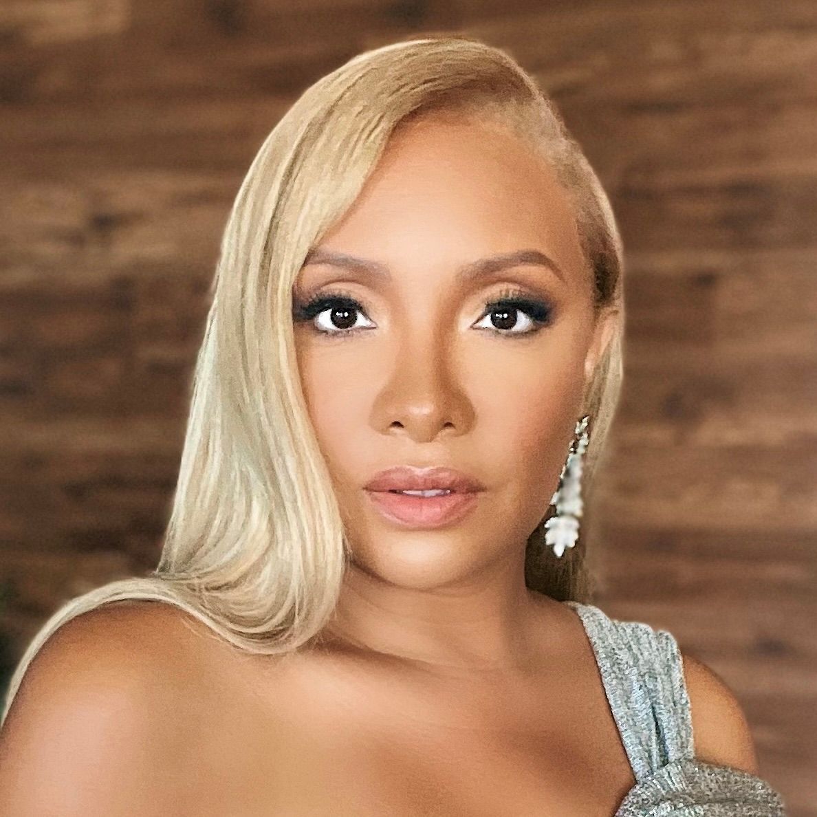 A person with long blonde hair and a light blue dress poses against a brown wooden backdrop.