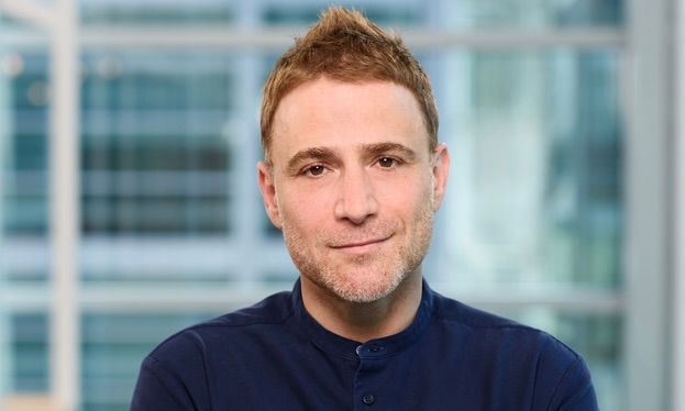 A person with short, spiky hair and light facial stubble wears a dark blue shirt against a blurred office background.