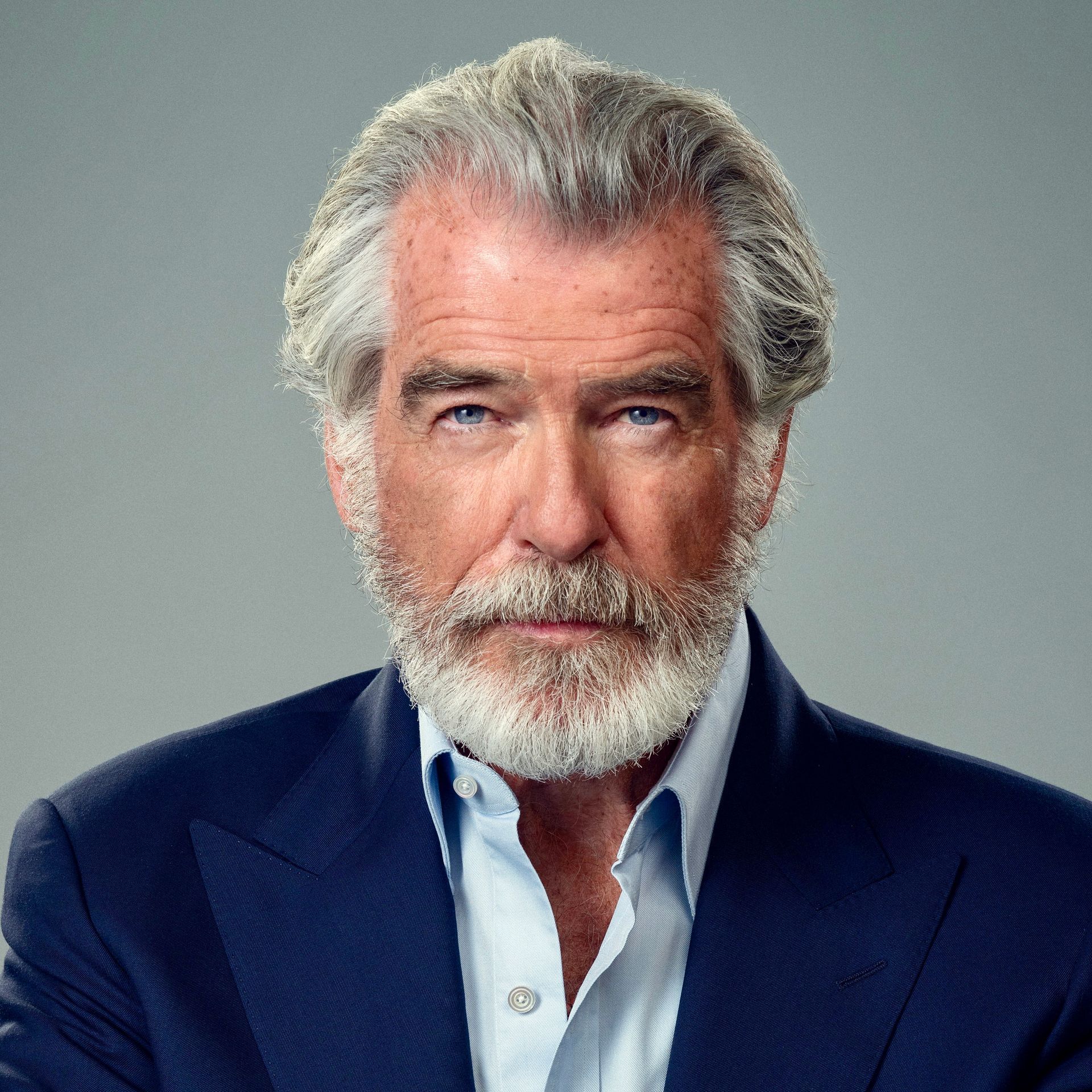 Headshot of a man with gray hair and a beard, wearing a navy blue suit jacket and light blue shirt against a gray background.