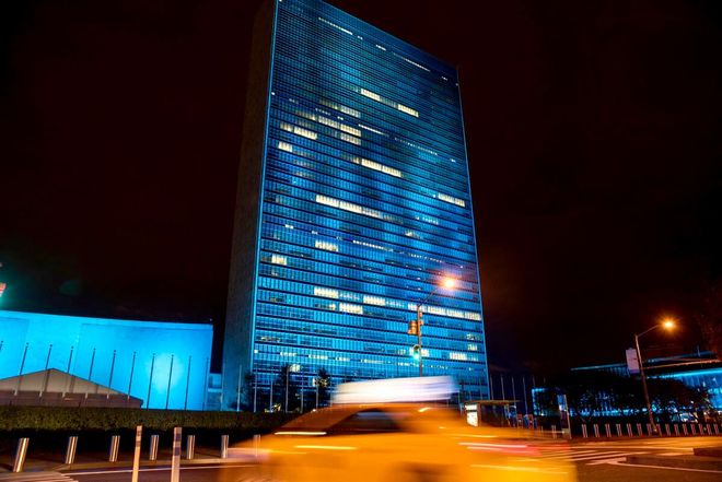 The United Nations Secretariat building in New York City illuminated in blue at night, with a motion-blurred taxi in front.