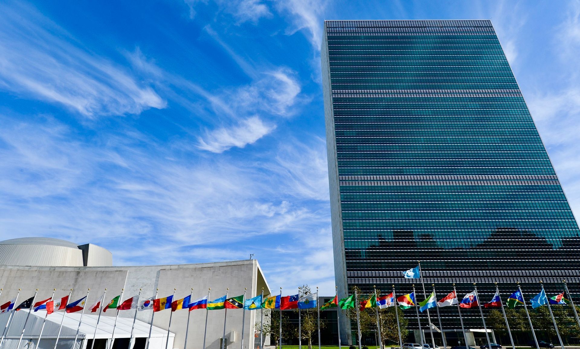 The United Nations Secretariat Building in New York City with a row of international flags under a blue, cloudy sky.