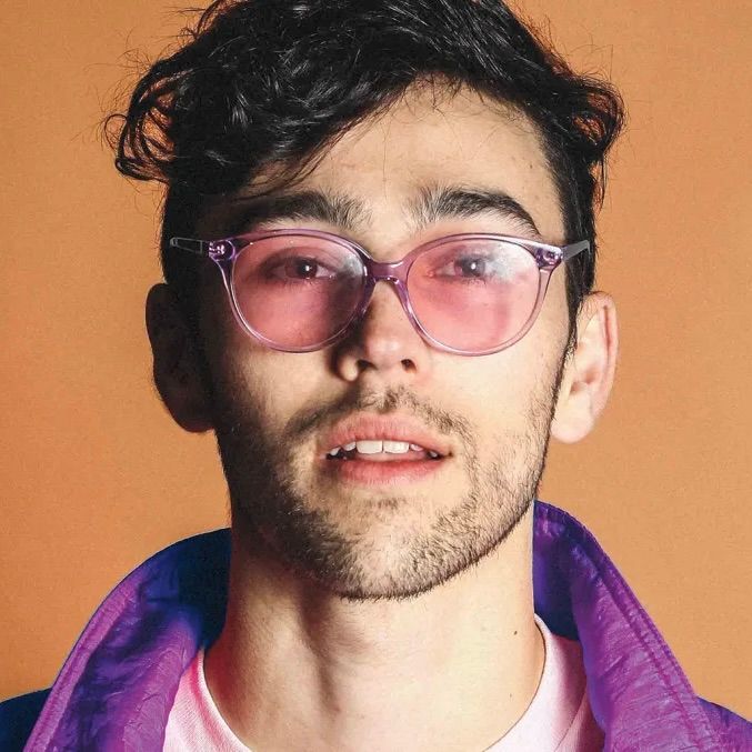 A person with dark, wavy hair wears translucent pink-tinted glasses and a purple jacket against a solid tan background.