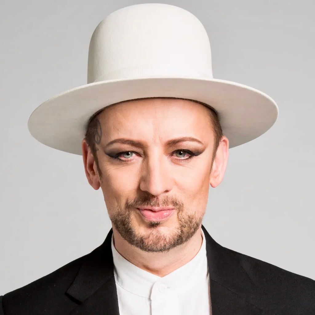 Boy George wearing a white wide-brimmed hat, dark eyeliner, and a black blazer over a white collared shirt.