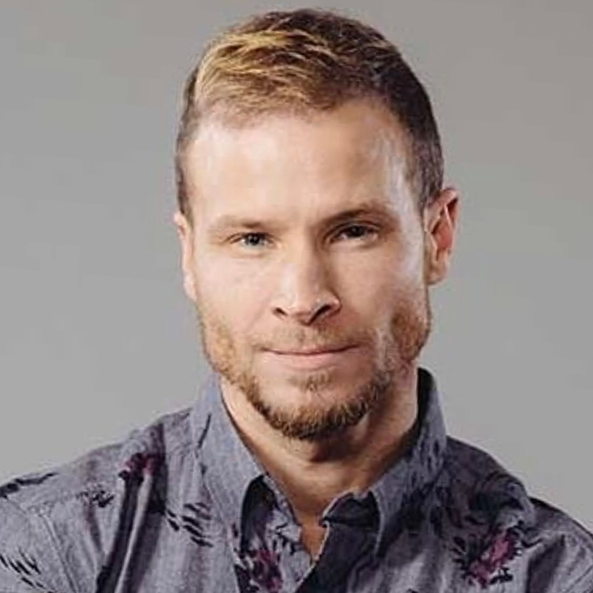 A person with short, light hair and a thin beard wears a gray patterned button-down shirt against a plain gray background.