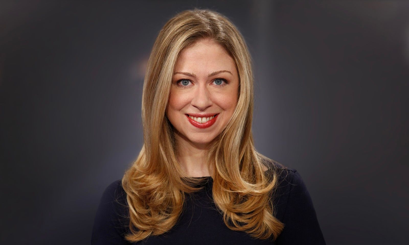 A person with shoulder-length blonde hair and a red-lipped smile, wearing a dark top against a plain gray background.