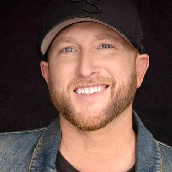 A man with a beard and a baseball cap smiles against a black background, wearing a denim jacket over a black shirt.