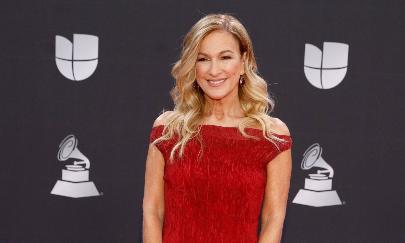 A smiling person in a red off-the-shoulder dress stands before a backdrop with Univision and Latin Grammy logos.