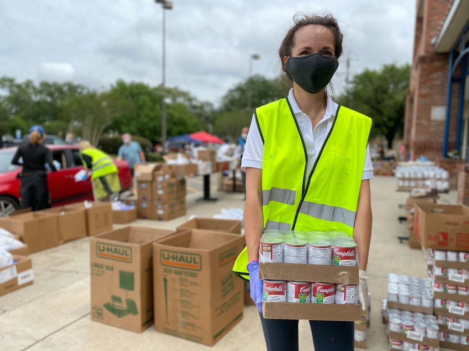 A volunteer in a yellow safety vest and face mask holds a tray of canned goods at an outdoor food distribution site.