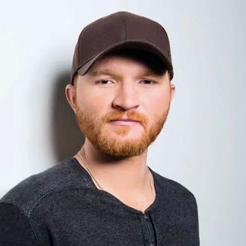 A person with a ginger beard wearing a dark baseball cap and a dark shirt against a plain light gray background.