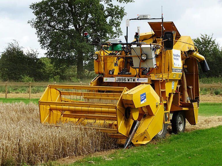 A yellow autonomous combine harvester operates in a field of wheat near a line of trees on a bright day.