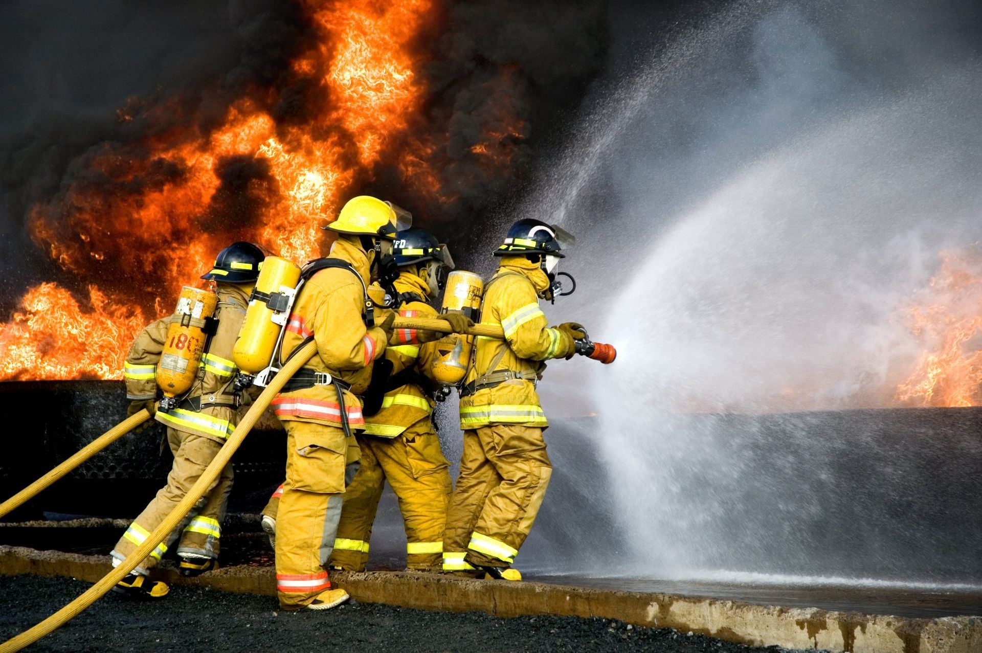 Four firefighters in yellow protective gear work together to aim a high-pressure hose at a large, intense structure fire.