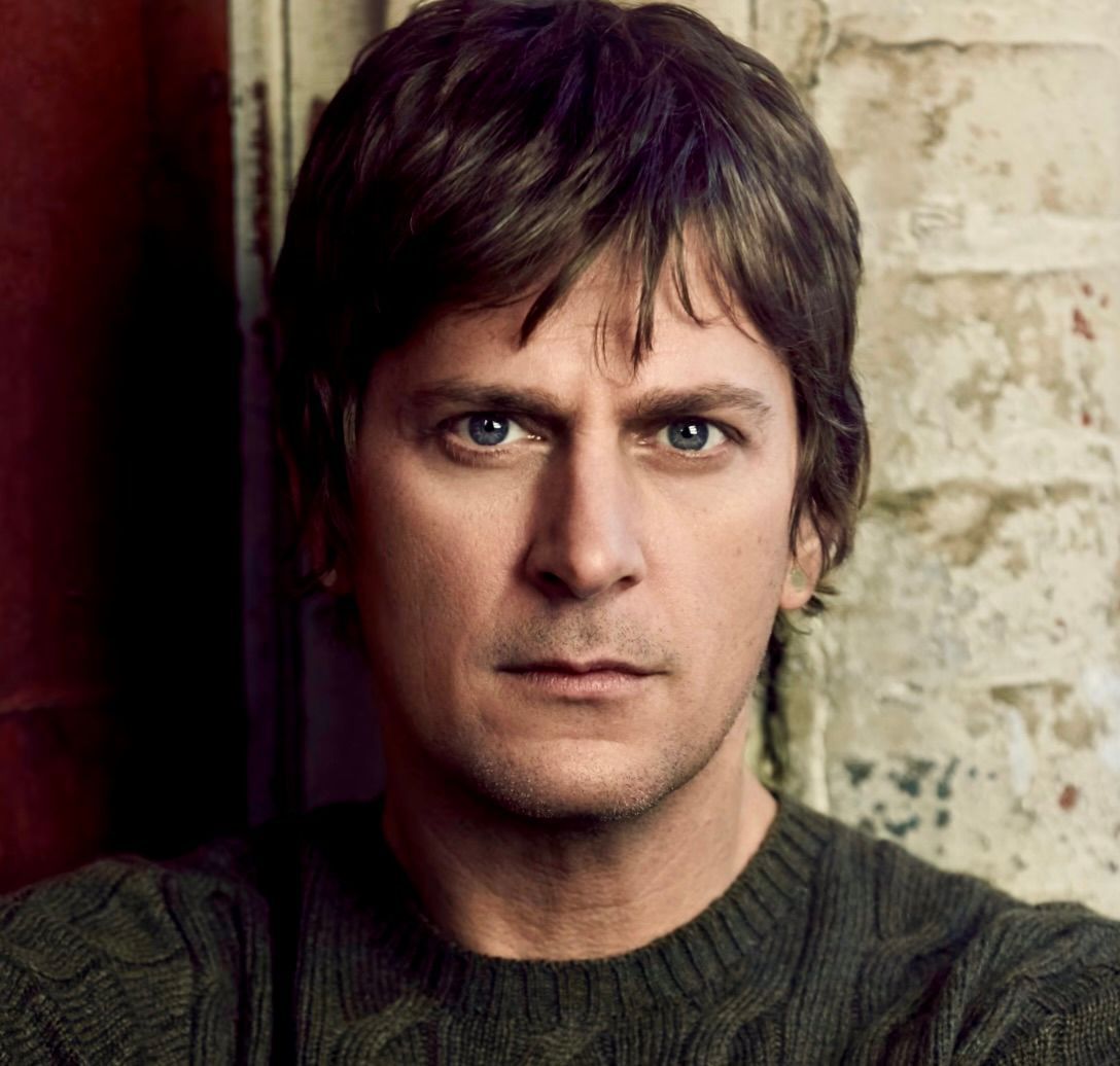 A person with dark hair and blue eyes wears a green cable-knit sweater, looking directly at the camera.