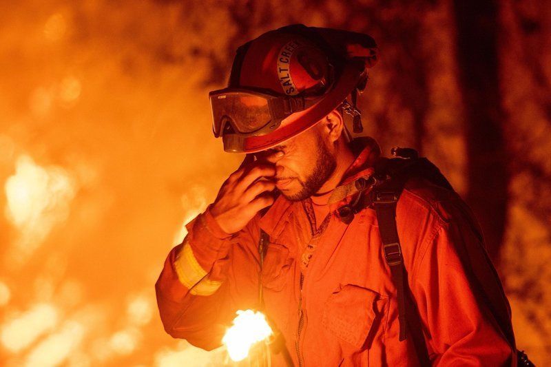 A firefighter in protective gear and goggles stands against a backdrop of intense, glowing flames.