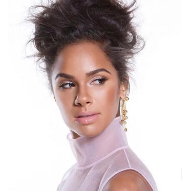 A person with voluminous dark hair and gold earrings, wearing a sheer high-neck top against a plain white background.