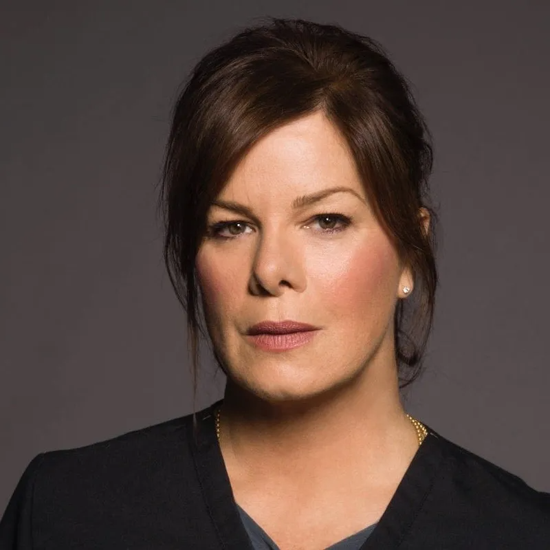 A person with brown hair pulled back, wearing a dark top, looking directly at the camera with a serious expression.