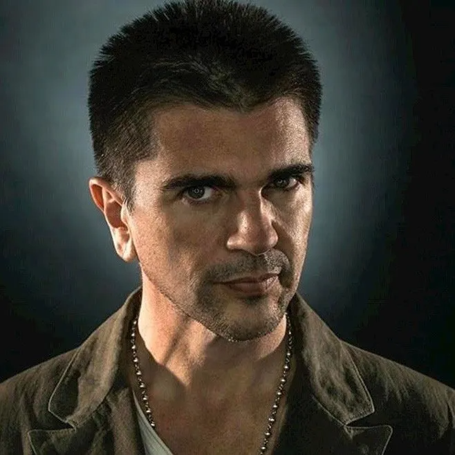 A person with short dark hair and a serious expression, wearing a jacket and beaded necklace against a dark background.