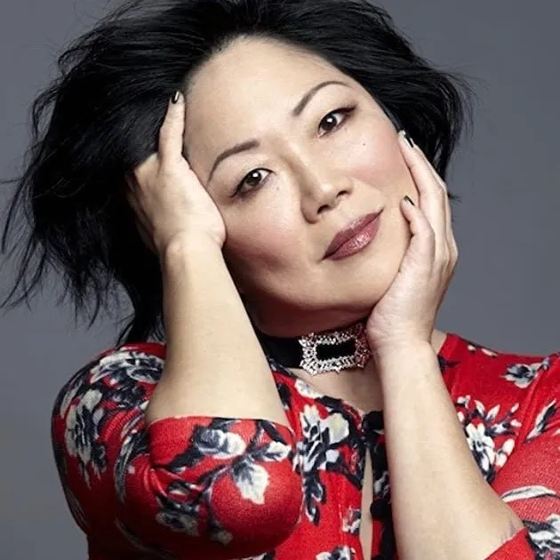 A person posing with both hands framing their face, wearing a red floral top and a black choker with a silver clasp.