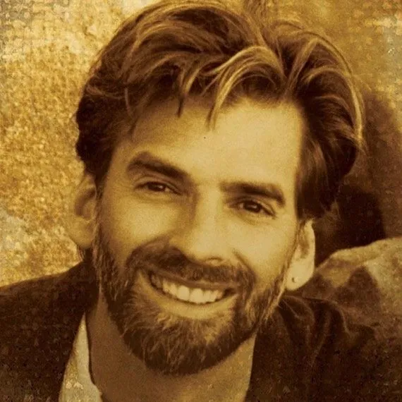 A sepia-toned headshot of a smiling person with a beard and dark, tousled hair, positioned against a textured backdrop.
