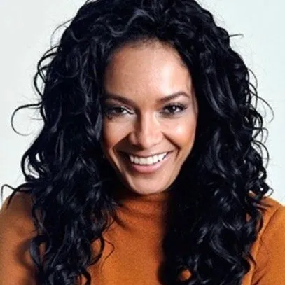 A person with long, dark, wavy hair smiles at the camera while wearing an orange-brown sweater against a white background.