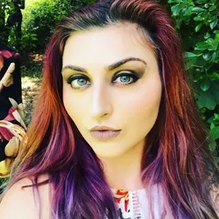 A person with shoulder-length, multi-colored hair fading from red to purple, wearing dark eye makeup in a garden setting.