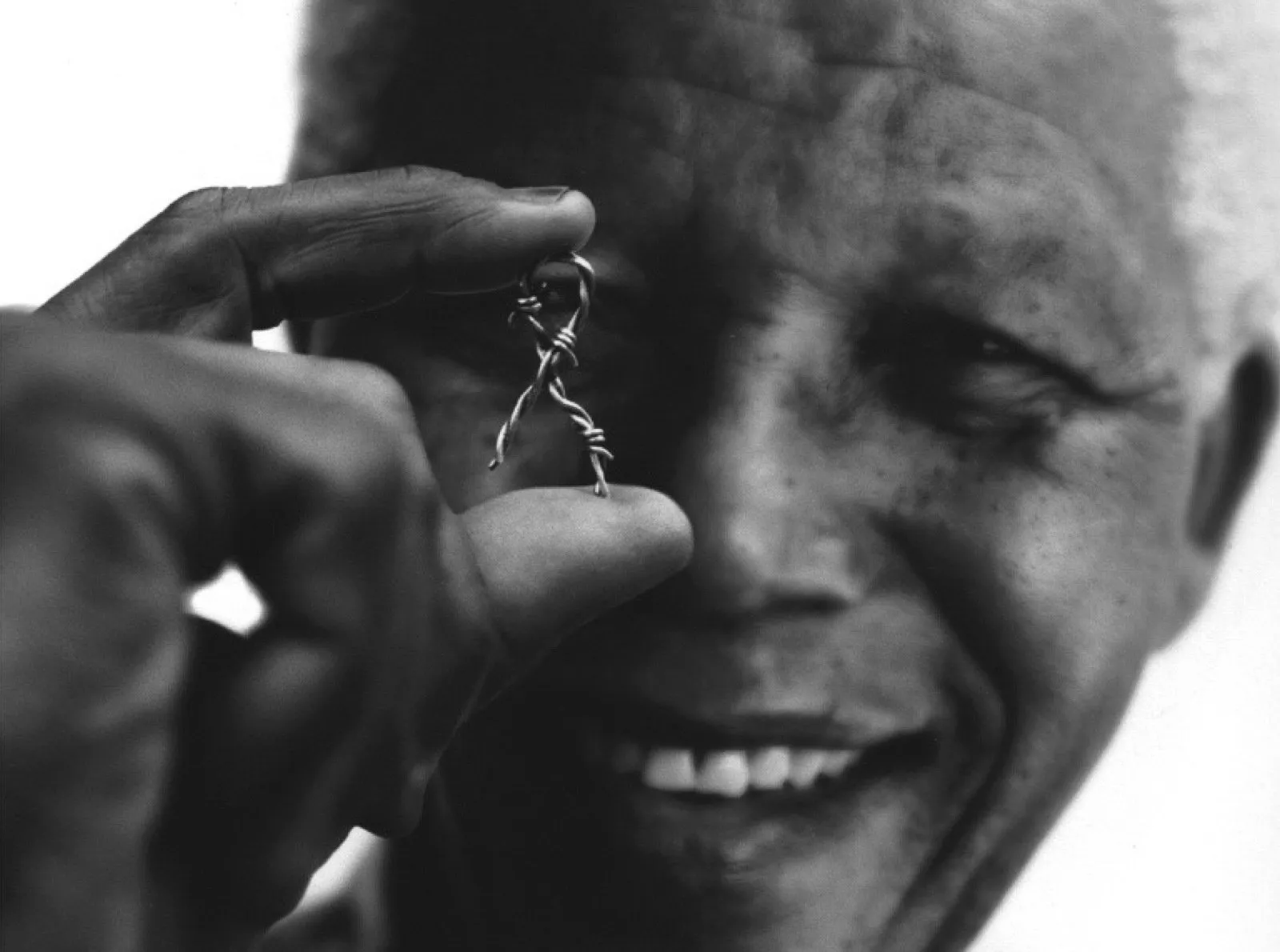 Nelson Mandela smiling while holding a small, barbed-wire AIDS awareness ribbon between his thumb and forefinger.