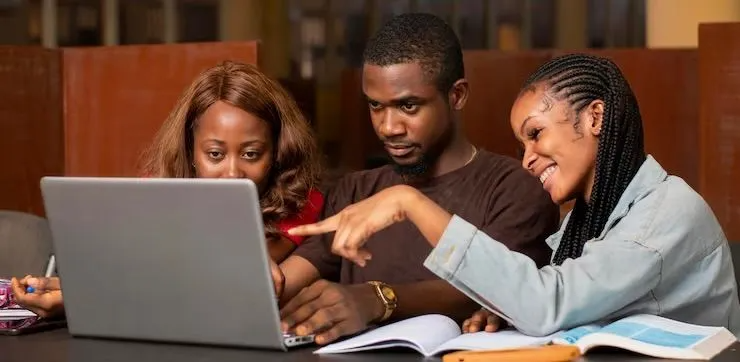 Three students collaborate at a desk, looking at a laptop screen while referring to open textbooks in a library setting.