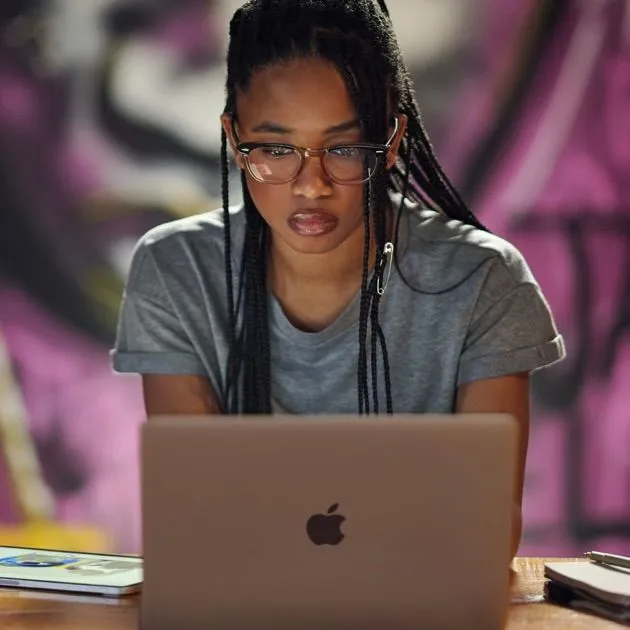 A focused person with braided hair and glasses works on an Apple laptop in a room with purple graffiti walls.