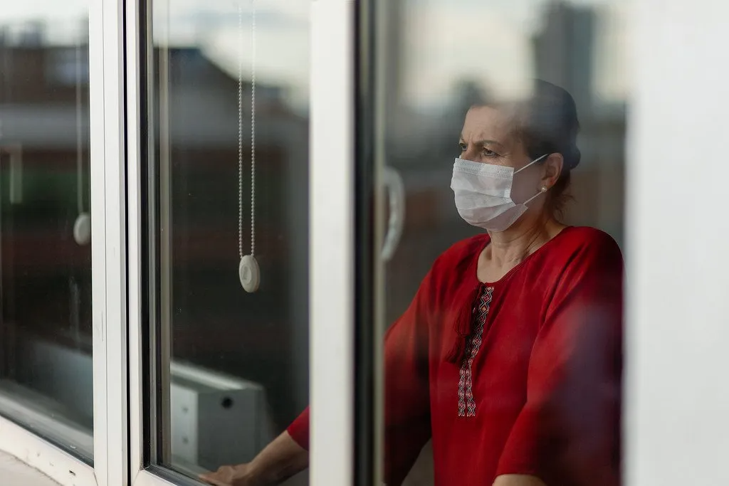 A person wearing a white face mask looking out through a window, wearing a red shirt.