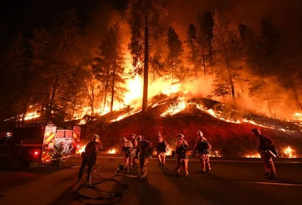 Firefighters stand on a dark road as a bright, intense wildfire burns through a forest on a hillside at night.