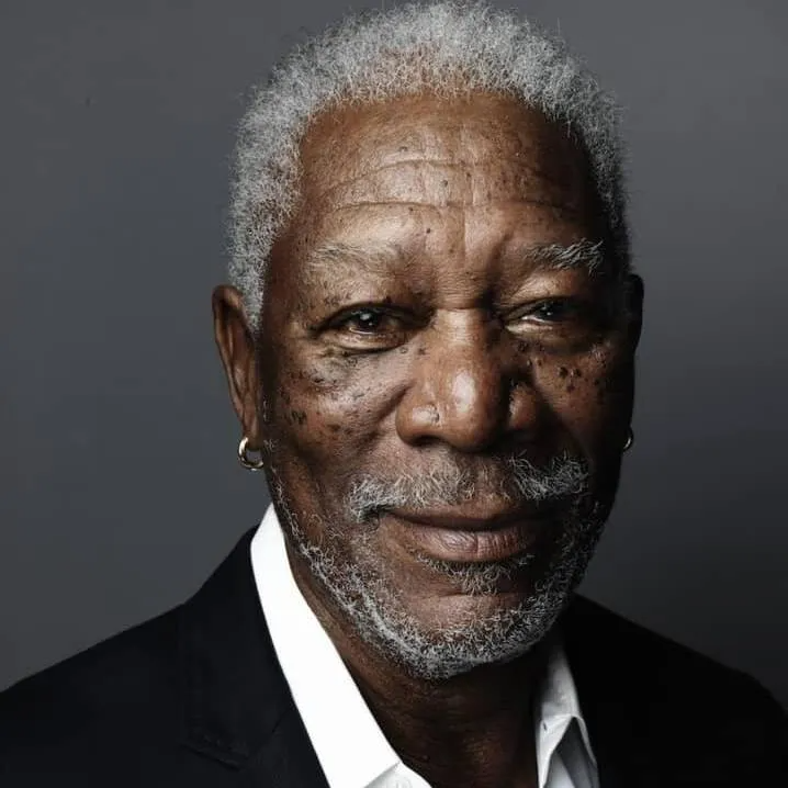 A headshot of Morgan Freeman against a grey background, smiling faintly while wearing a suit jacket and small hoop earring.