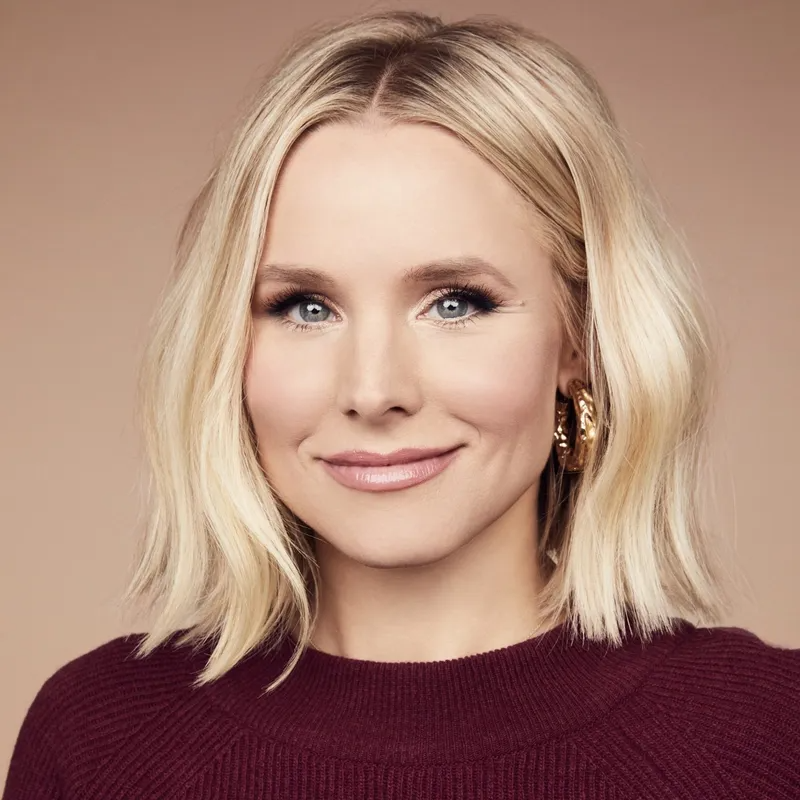 Portrait of a blonde woman with a shoulder-length bob, wearing a dark red sweater against a neutral, light brown background.