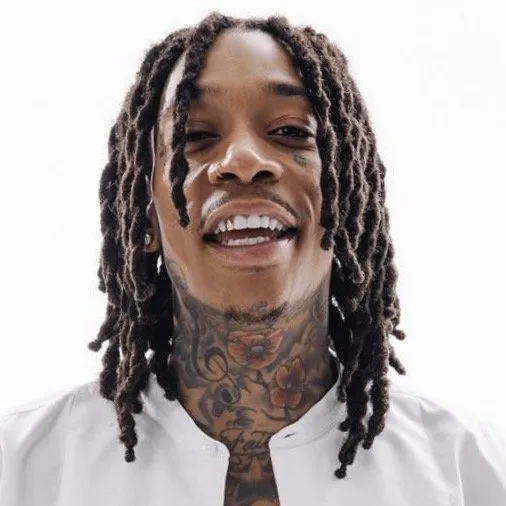 A smiling person with shoulder-length dreadlocks and extensive neck tattoos, wearing a white shirt against a white backdrop.