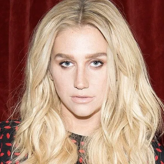 Kesha with long, blonde, wavy hair against a red curtain background, wearing a black patterned top.