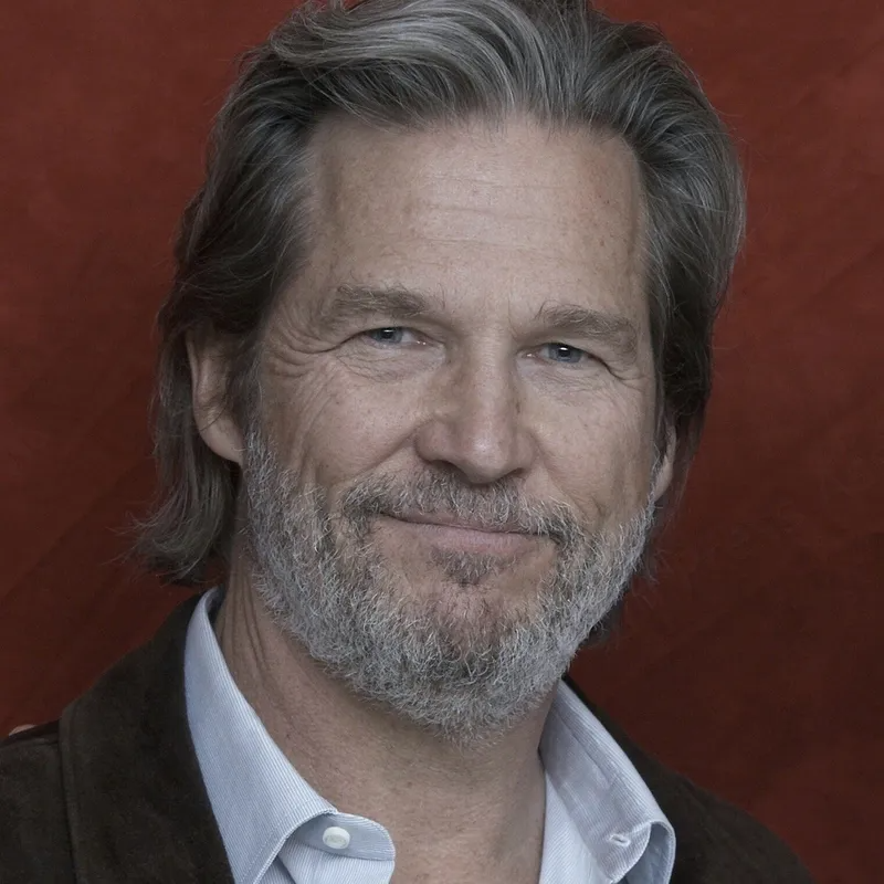 A headshot of actor Jeff Bridges smiling warmly, wearing a white shirt and dark jacket against a dark red background.