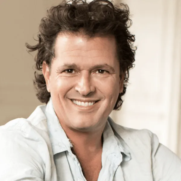 A person with dark, wavy hair, wearing a light-colored button-down shirt, smiles warmly in a bright, indoor setting.