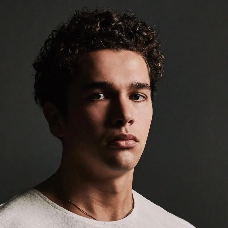 Headshot of a person with curly dark hair against a dark gray background, wearing a white shirt and a thin necklace.
