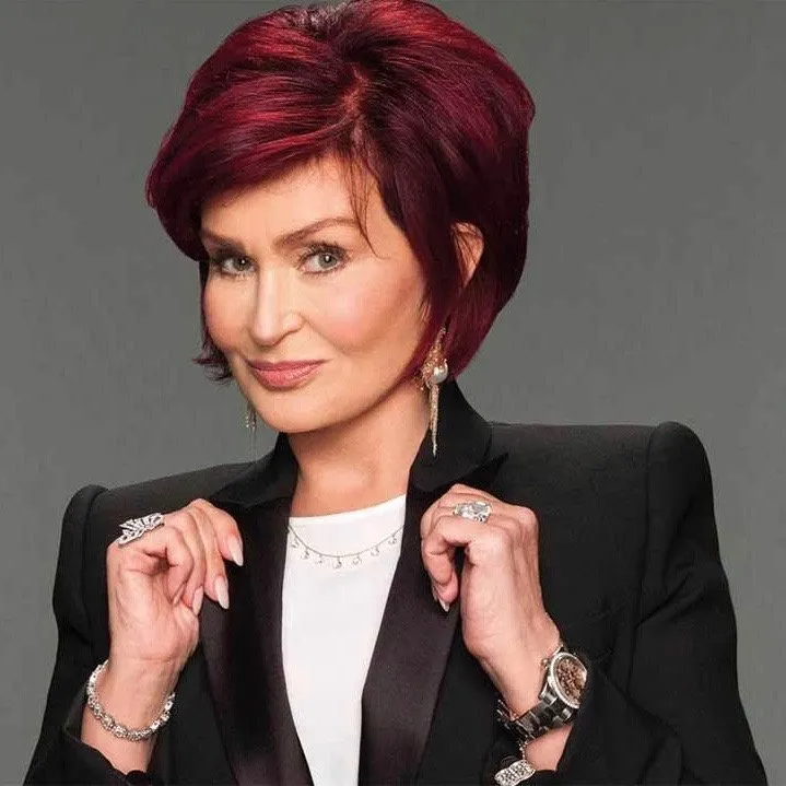 Sharon Osbourne with signature red hair, wearing a black blazer and white top, smiling while holding her lapels.