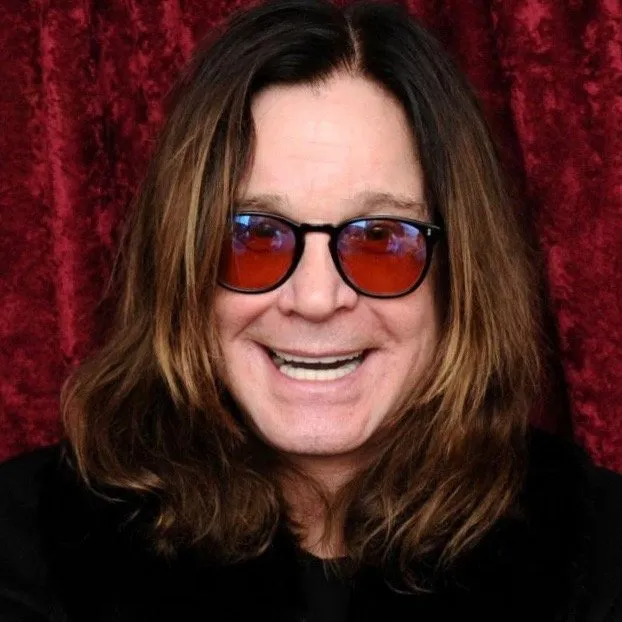 Ozzy Osbourne smiling at the camera, wearing round, reddish-tinted glasses in front of a red, textured curtain background.
