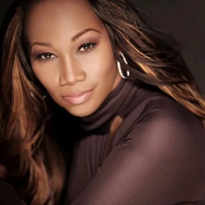 A portrait of a person with long, highlighted brown hair wearing a dark turtleneck and hoop earrings against a dark background.