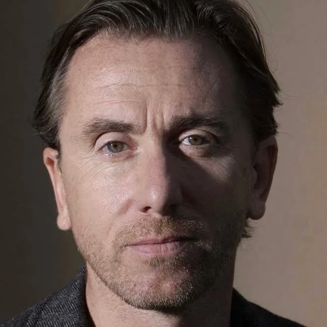 Close-up portrait of Tim Roth, facing forward with a neutral expression, against a plain, dimly lit background.