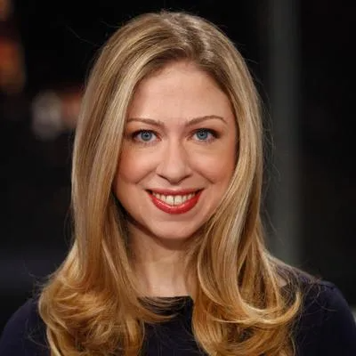 A smiling person with long blonde hair and blue eyes against a dark, out-of-focus background.