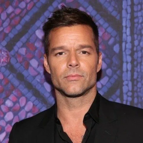 Ricky Martin wearing a black shirt in front of a purple mosaic background, looking directly at the camera.