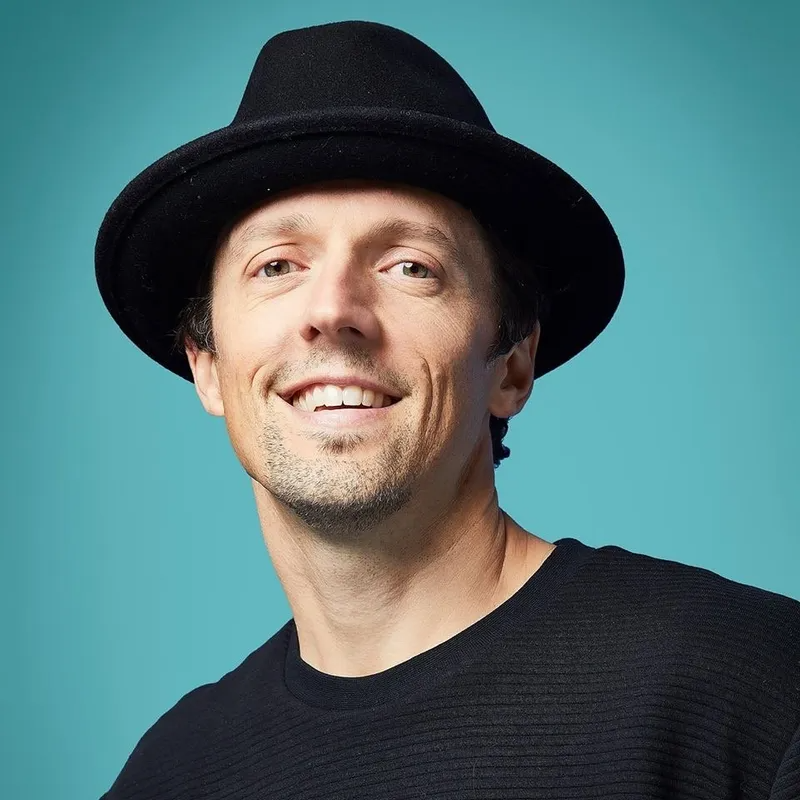 A person wearing a black fedora and a black shirt smiles at the camera against a solid light blue background.