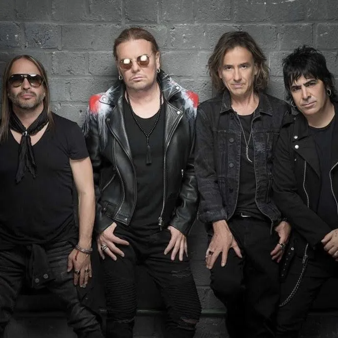 Four men in black clothing and sunglasses posing together against a gray brick wall.