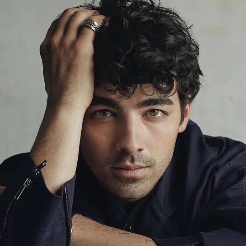 A portrait of a person with dark, curly hair and light eyes, resting their head on their hand against a plain background.